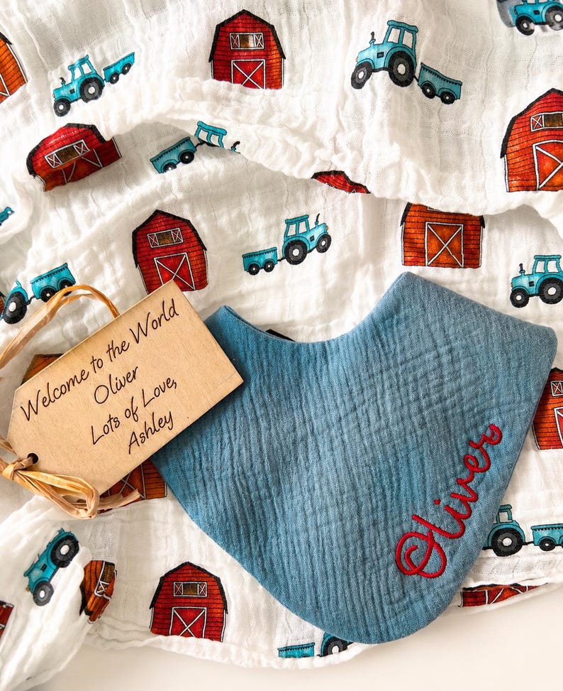 May include: A blue muslin bandana bib with the name Oliver embroidered in red thread, placed on a white muslin blanket featuring a repeating pattern of red barns and blue tractors, accompanied by a rectangular wooden tag reading Welcome to the World Oliver Lots of Love, Ashley.