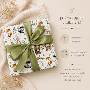 May include: A square gift box wrapped in white paper featuring a pattern of woodland animals including foxes, raccoons, rabbits, and deer with green foliage. The box is tied with a wide olive green satin ribbon bow. Text reads gift wrapping available $5.