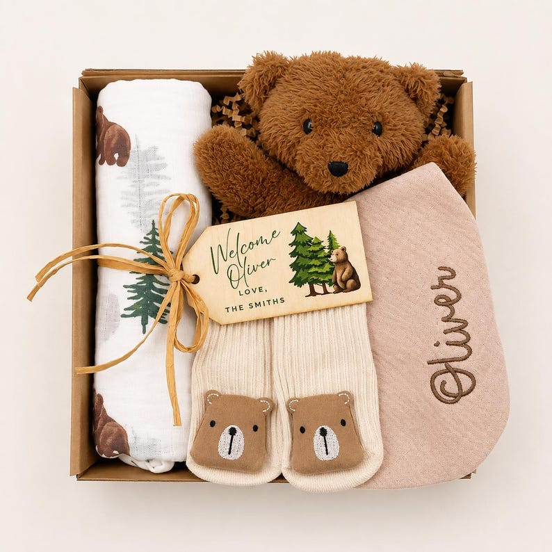 May include: A gift box containing a brown plush teddy bear, a white swaddle blanket with bear and tree prints, a pair of cream ribbed socks with bear faces, and a pink bib embroidered with the name Oliver. A wooden tag reads Welcome Oliver Love, The Smiths.