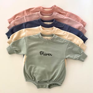May include: A stack of five baby rompers in various colors: dusty rose, navy blue, beige, and sage green. The sage green romper has the name "Oliver" embroidered on the front. Each romper has snap closures and a crew neck.