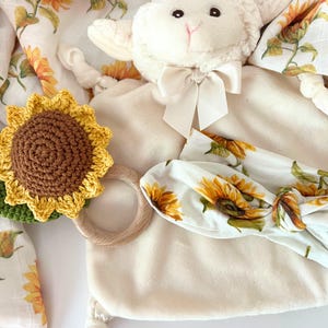 May include: A white plush lamb toy with a brown crocheted sunflower rattle, a wooden teething ring, and a white headband with a sunflower print.