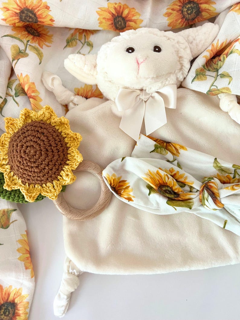 May include: A white plush lamb toy with a brown crocheted sunflower rattle, a wooden teething ring, and a white headband with a sunflower print.