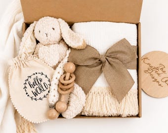 May include: A gift box containing a cream-colored plush bunny, a white muslin blanket with a large brown linen bow and fringe trim, a wooden rattle, and a cream fabric pouch with the text hello WORLD printed inside a circular leaf wreath.