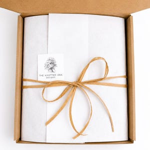May include: A brown cardboard shipping box containing white tissue paper wrapped around an item. The paper is tied with a tan raffia ribbon and features a square white sticker with a tree illustration and the text The Knotted Oak Designs.