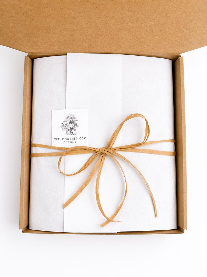 May include: A brown cardboard shipping box containing white tissue paper wrapped around an item. The paper is tied with a tan raffia ribbon and features a square white sticker with a tree illustration and the text The Knotted Oak Designs.