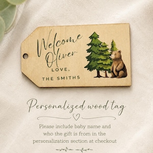 May include: A rectangular wooden gift tag with a hole on the left side. The tag features a printed illustration of green pine trees and a brown bear. The text reads Welcome Oliver LOVE, THE SMITHS. The tag has a light wood grain finish.