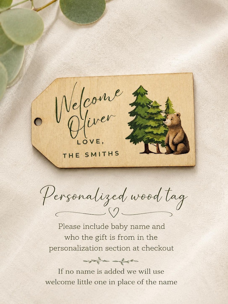 May include: A rectangular wooden gift tag with a hole on the left side. The tag features a printed illustration of green pine trees and a brown bear. The text reads Welcome Oliver LOVE, THE SMITHS. The tag has a light wood grain finish.