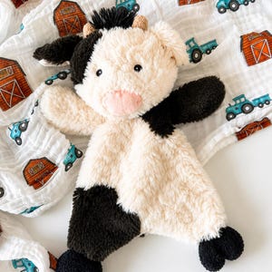 May include: A plush cow security blanket featuring a soft cream and black faux fur body, a pink snout, and small horns. The toy is placed on a white muslin fabric patterned with small red barns and blue tractors, creating a farm-themed design.