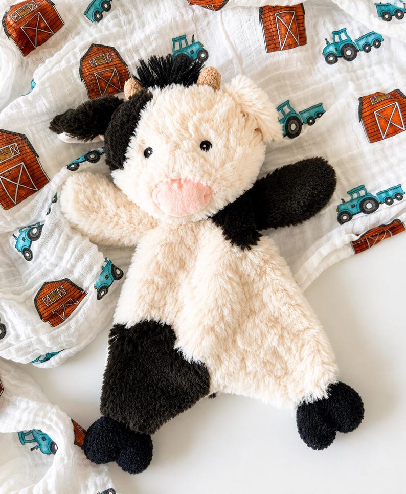 May include: A plush cow security blanket featuring a soft cream and black faux fur body, a pink snout, and small horns. The toy is placed on a white muslin fabric patterned with small red barns and blue tractors, creating a farm-themed design.