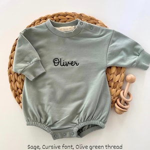 Puede incluir: Body de beb&eacute; verde salvia con el nombre "Oliver" bordado en cursiva con hilo verde oliva. El body tiene mangas largas y cierres de bot&oacute;n a presi&oacute;n. Un sonajero de madera est&aacute; al lado.