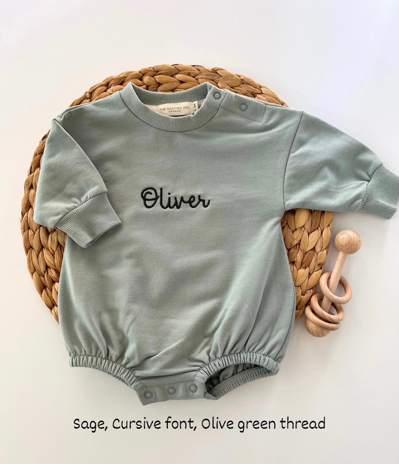 Puede incluir: Body de beb&eacute; verde salvia con el nombre "Oliver" bordado en cursiva con hilo verde oliva. El body tiene mangas largas y cierres de bot&oacute;n a presi&oacute;n. Un sonajero de madera est&aacute; al lado.