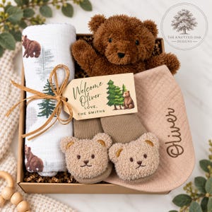 May include: A gift box containing a white swaddle blanket with brown bear and green tree prints, a brown plush teddy bear, a tan bib embroidered with the name Oliver, and tan baby booties with bear faces. A wooden tag reads Welcome Oliver.