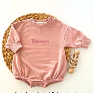 Puede incluir: Un pelele de beb&eacute; color peon&iacute;a con mangas largas y cuello redondo. El nombre "Emerson" est&aacute; bordado con hilo fucsia. El pelele est&aacute; sobre una estera tejida con un juguete de madera.