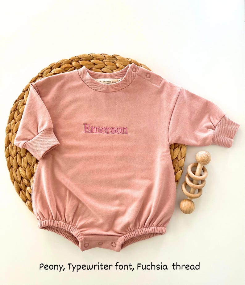Puede incluir: Un pelele de beb&eacute; color peon&iacute;a con mangas largas y cuello redondo. El nombre "Emerson" est&aacute; bordado con hilo fucsia. El pelele est&aacute; sobre una estera tejida con un juguete de madera.