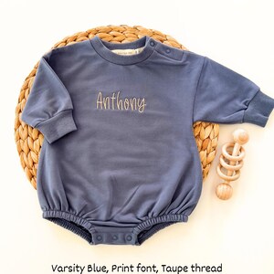Puede incluir: Body de beb&eacute; azul marino con el nombre "Anthony" bordado en una fuente de impresi&oacute;n con hilo color taupe. El body de manga larga tiene cuello redondo y cierres de bot&oacute;n a presi&oacute;n. Un juguete de madera est&aacute; al lado.