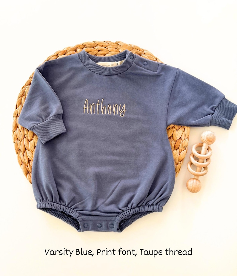 Puede incluir: Body de beb&eacute; azul marino con el nombre "Anthony" bordado en una fuente de impresi&oacute;n con hilo color taupe. El body de manga larga tiene cuello redondo y cierres de bot&oacute;n a presi&oacute;n. Un juguete de madera est&aacute; al lado.