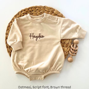 Puede incluir: Body de beb&eacute; color avena con el nombre "Hayden" bordado en fuente cursiva marr&oacute;n. El body de manga larga tiene cierres de bot&oacute;n a presi&oacute;n en la parte inferior y en el hombro. Un sonajero de madera est&aacute; junto al body sobre una estera tejida.