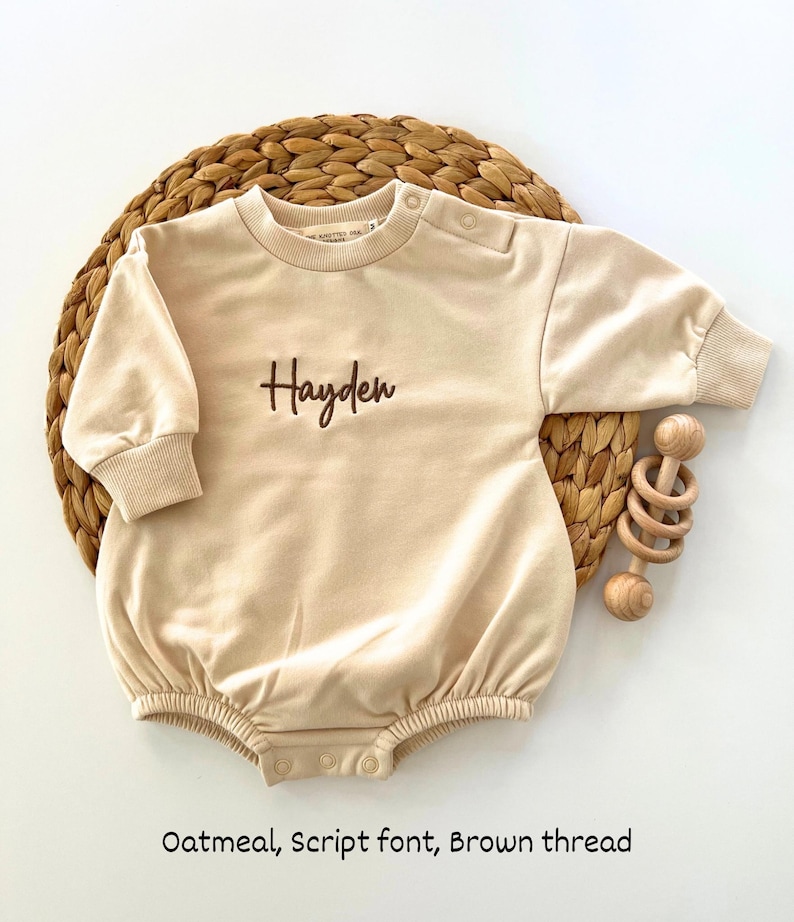 Puede incluir: Body de beb&eacute; color avena con el nombre "Hayden" bordado en fuente cursiva marr&oacute;n. El body de manga larga tiene cierres de bot&oacute;n a presi&oacute;n en la parte inferior y en el hombro. Un sonajero de madera est&aacute; junto al body sobre una estera tejida.