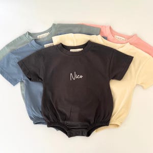 May include: A collection of baby rompers in various pastel colors, including blue, green, yellow, and pink. The dark gray romper in the center features the name "Nico" in white script. Each romper has short sleeves and a round neckline.