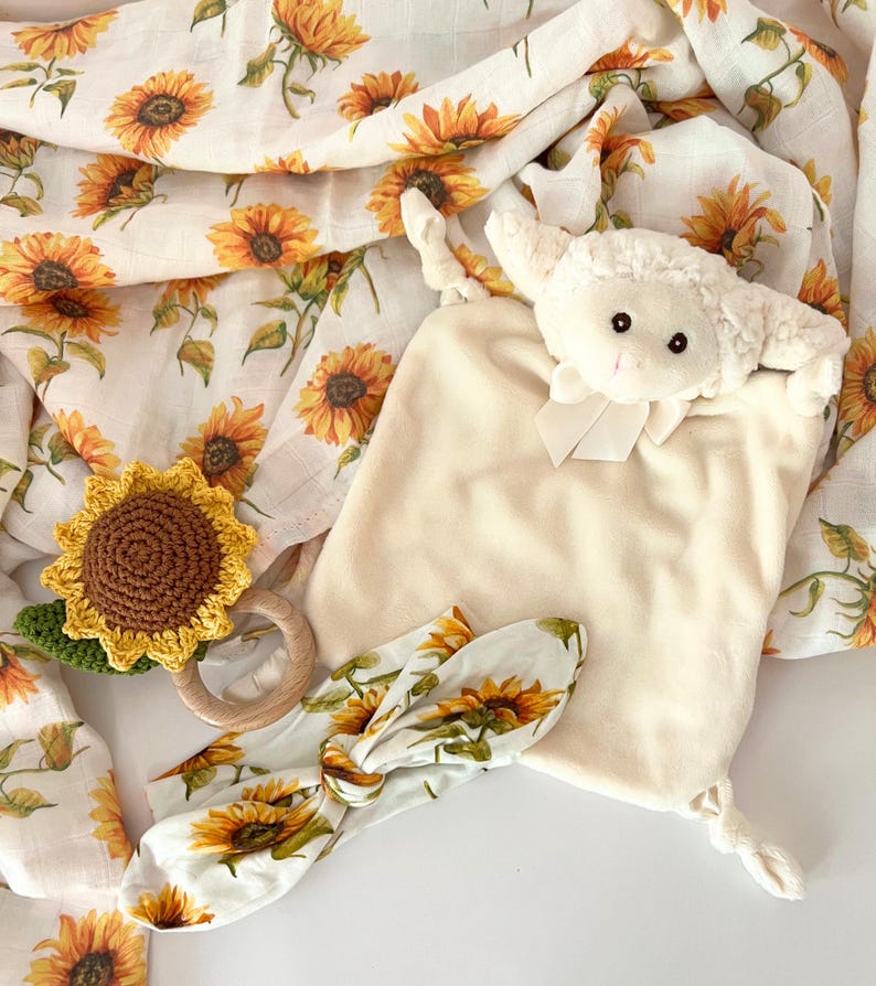 May include: A white plush lamb toy with a sunflower print headband, a crocheted sunflower rattle, and a white muslin swaddle blanket with a sunflower print.