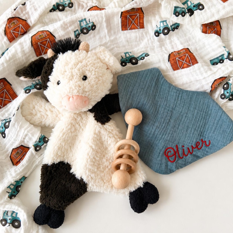 May include: A plush cow security blanket with black and white fur, a blue muslin swaddle blanket featuring a farm tractor and barn print, a wooden rattle, and a blue triangular bib embroidered with the name Oliver in red cursive script.