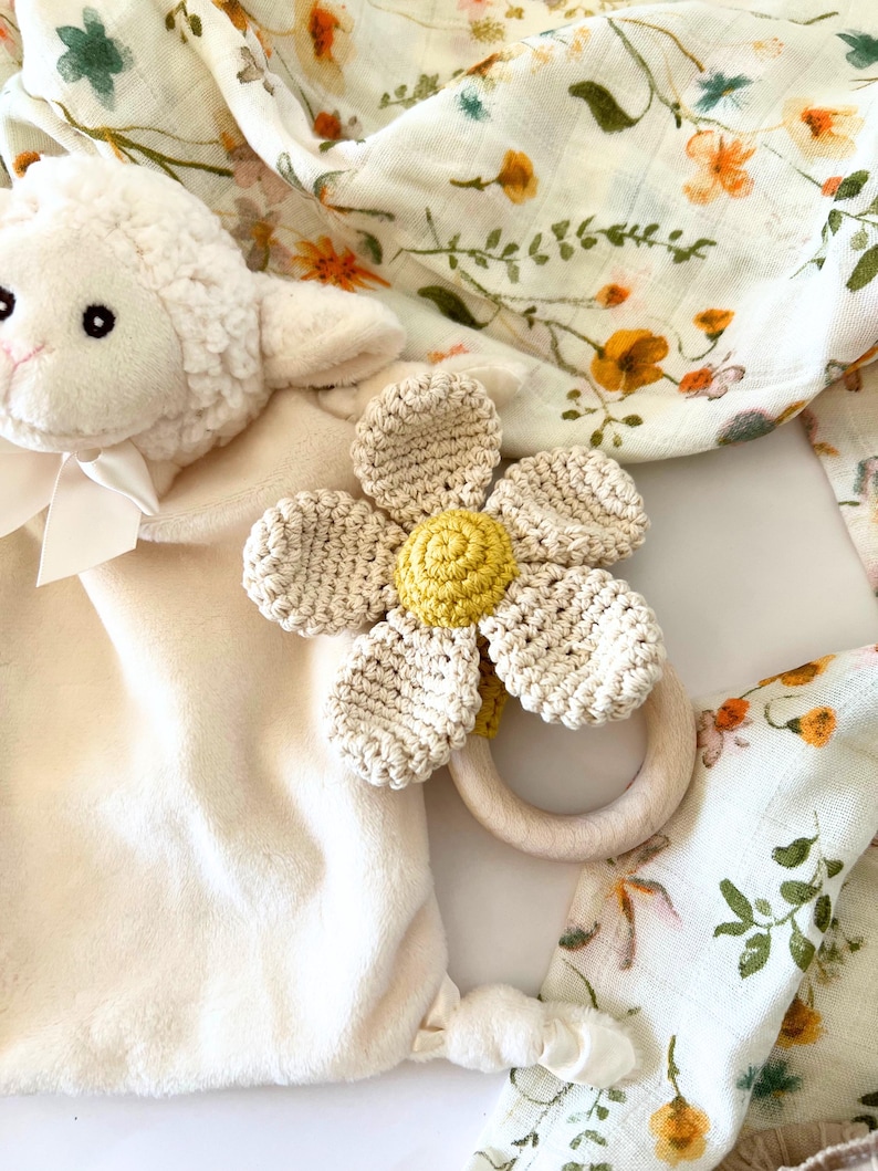 May include: A crocheted flower teether with a wooden ring, a plush white lamb toy, and a floral muslin swaddle blanket.