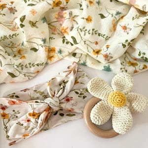 May include: A set of baby items featuring a floral print. The set includes a swaddle blanket, a headband, and a crocheted flower teether.