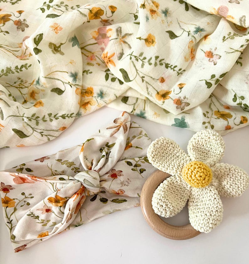 May include: A set of baby items featuring a floral print. The set includes a swaddle blanket, a headband, and a crocheted flower teether.