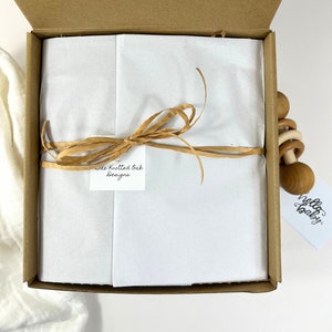 May include: A brown cardboard gift box with a white tissue paper lining. The box is tied with a brown ribbon and has a tag that says "The Knotted Oak Designs". A wooden baby rattle with a "hello baby" tag is visible on the right side of the box.