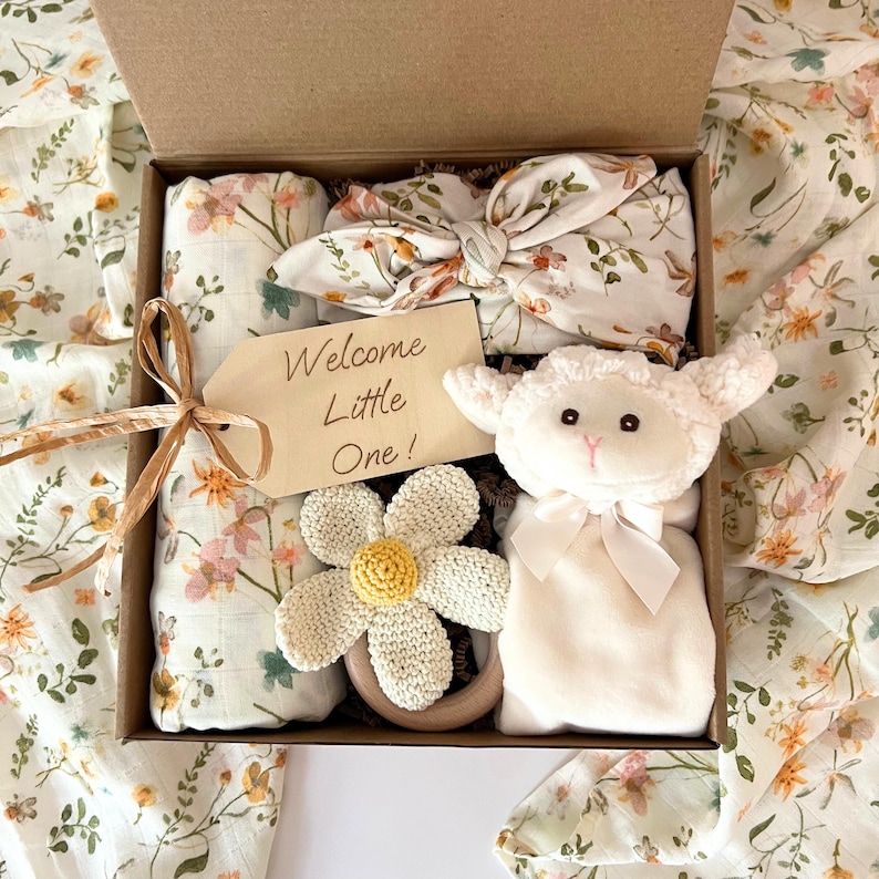 May include: A gift box filled with baby items, including a floral print swaddle blanket, a crocheted flower teether, a plush lamb toy, and a floral print headband. The box also includes a wooden tag that says "Welcome Little One!"