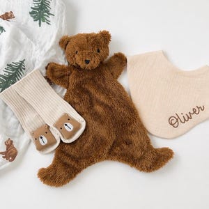 May include: A brown plush bear security blanket, a pair of cream-coloured ribbed socks with bear face patches, and a beige muslin bib embroidered with the name Oliver, alongside a white fabric featuring a green pine tree and brown bear pattern.