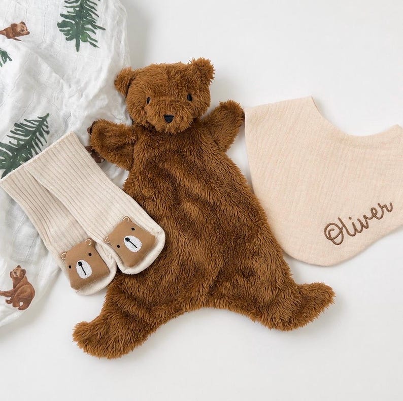 May include: A brown plush bear security blanket, a pair of cream-coloured ribbed socks with bear face patches, and a beige muslin bib embroidered with the name Oliver, alongside a white fabric featuring a green pine tree and brown bear pattern.