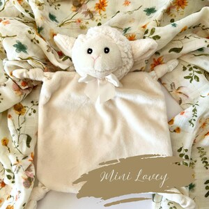 May include: A white plush lamb lovey with a satin bow. The lovey is lying on a white and green floral muslin swaddle blanket. The text "Mini Lovey" is written on a brown background.