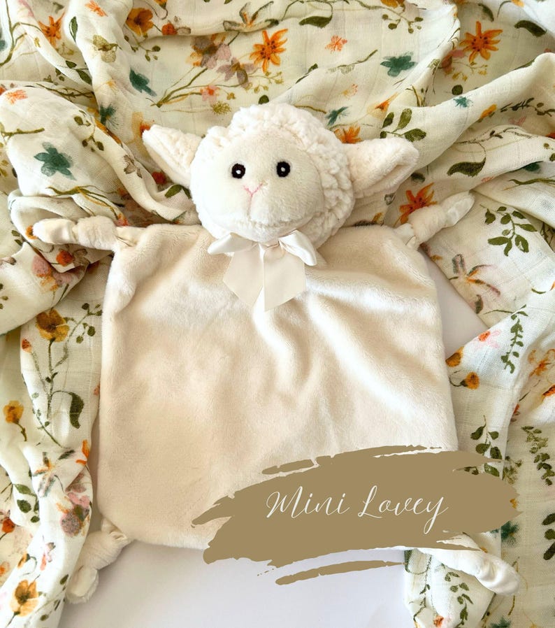 May include: A white plush lamb lovey with a satin bow. The lovey is lying on a white and green floral muslin swaddle blanket. The text "Mini Lovey" is written on a brown background.