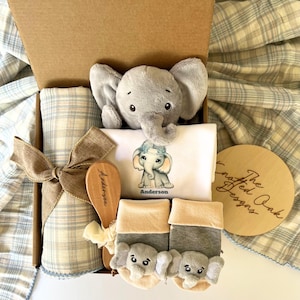 May include: A baby gift set in a cardboard box, featuring a gray plush elephant, a white onesie with an elephant graphic, a wooden brush, and a pair of elephant-themed socks. A plaid blanket and a wooden disc are also included.