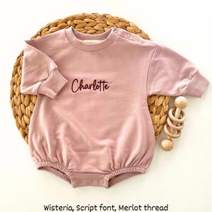Puede incluir: Un body de beb&eacute; rosa claro con el nombre "Charlotte" bordado en letra cursiva con hilo Merlot. El body tiene mangas largas y cuello redondo. Un juguete de madera est&aacute; al lado del body.