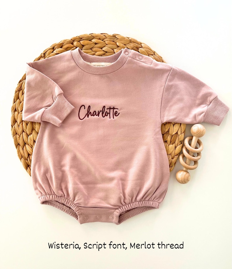 Puede incluir: Un body de beb&eacute; rosa claro con el nombre "Charlotte" bordado en letra cursiva con hilo Merlot. El body tiene mangas largas y cuello redondo. Un juguete de madera est&aacute; al lado del body.