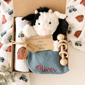 May include: A gift box containing a plush black and white cow, a white muslin swaddle with red barn and blue tractor prints, a blue fabric bib embroidered with the name Oliver in red, and a wooden rattle. A tag reads: Welcome to the World Oliver Lots of Love, Ashley.