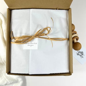 K&ouml;nnte beinhalten: Ein brauner Karton mit einer wei&szlig;en Seidenpapierauskleidung. Die Schachtel ist mit einem braunen Band gebunden und hat ein Etikett mit der Aufschrift "The Knotted Oak Designs". Rechts neben der Schachtel ist ein h&ouml;lzerner Babyrassel mit einem Etikett mit der Aufschrift "hello baby" zu sehen.