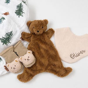 May include: A brown plush bear security blanket, a pair of tan baby socks with small bear faces, and a beige fabric bib embroidered with the name Oliver, alongside a white muslin swaddle blanket featuring a green pine tree pattern.