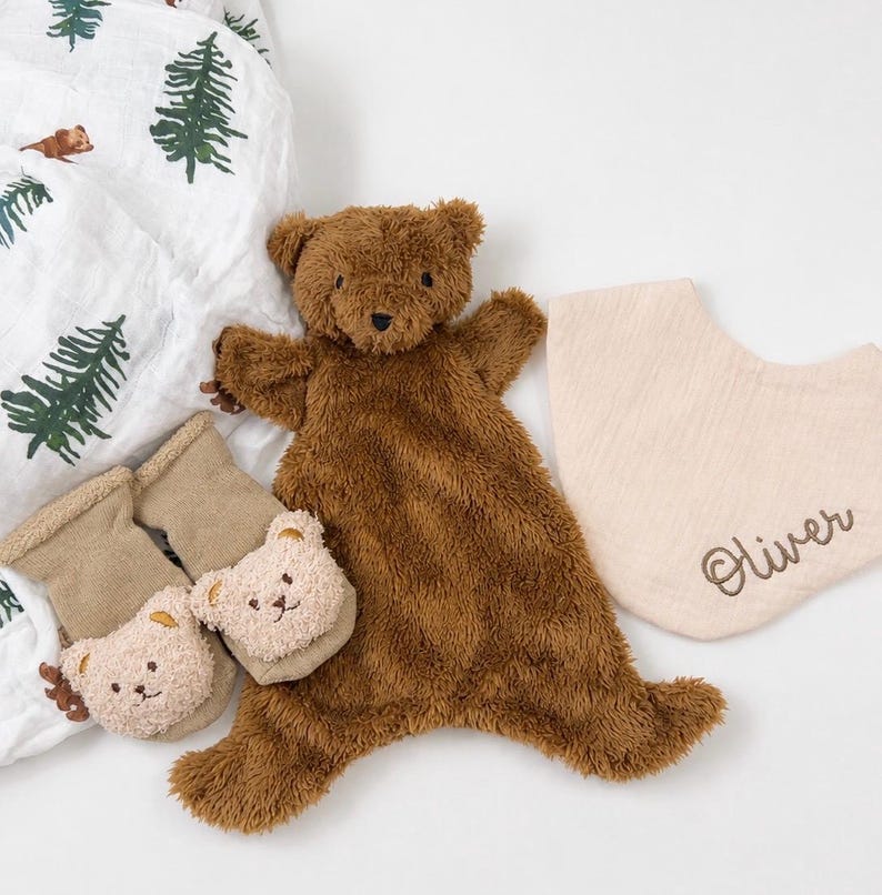 May include: A brown plush bear security blanket, a pair of tan baby socks with small bear faces, and a beige fabric bib embroidered with the name Oliver, alongside a white muslin swaddle blanket featuring a green pine tree pattern.