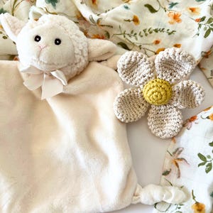 May include: A white plush lamb security blanket with a crocheted flower attached. The flower is beige with a yellow center. The blanket is made of soft, cuddly fabric and is perfect for babies and toddlers.
