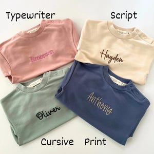 Puede incluir: Cuatro sudaderas para beb&eacute;s en rosa, crema, verde salvia y azul, cada una bordada con un nombre en una fuente diferente. El texto "Font choice", "Typewriter", "Script", "Cursive" y "Print" es visible. Tallas disponibles: 0-3, 3-6 y 6-12 meses.