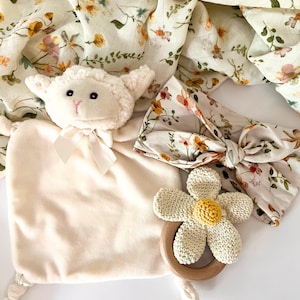 May include: A baby gift set with a plush white lamb, a floral headband, a crocheted flower teether, and a muslin swaddle blanket with a floral print.