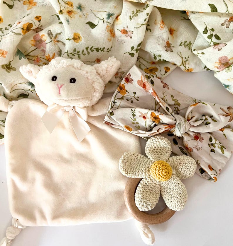 May include: A baby gift set with a plush white lamb, a floral headband, a crocheted flower teether, and a muslin swaddle blanket with a floral print.