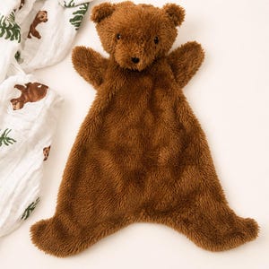 May include: A brown plush bear security blanket with a soft, shaggy texture. The bear features a rounded head with small ears, dark eyes, and a stitched nose. The body is flat and wide, extending into four points, resembling a small blanket.