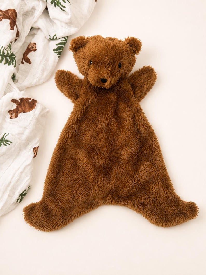 May include: A brown plush bear security blanket with a soft, shaggy texture. The bear features a rounded head with small ears, dark eyes, and a stitched nose. The body is flat and wide, extending into four points, resembling a small blanket.