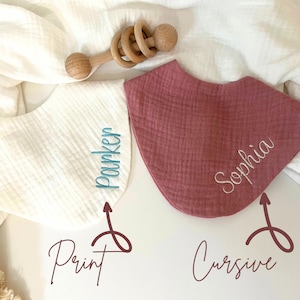 May include: Two baby bibs, one white and one mauve, with embroidered names. The white bib has "Parker" in print, and the mauve bib has "Sophia" in cursive. A wooden baby rattle rests above the bibs.