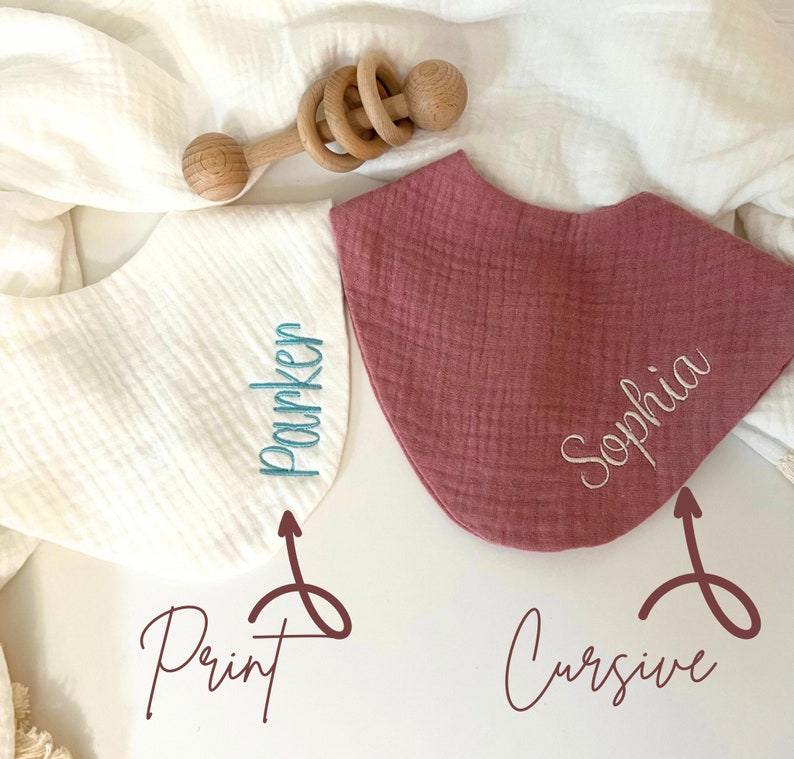 May include: Two baby bibs, one white and one mauve, with embroidered names. The white bib has "Parker" in print, and the mauve bib has "Sophia" in cursive. A wooden baby rattle rests above the bibs.