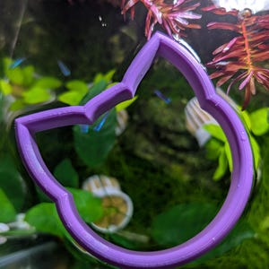 May include: A purple, cat-shaped cookie cutter is partially submerged in water, surrounded by green aquatic plants and red-purple stems. Small blue fish swim in the background. The cutter's design is a stylized cat head with pointed ears.